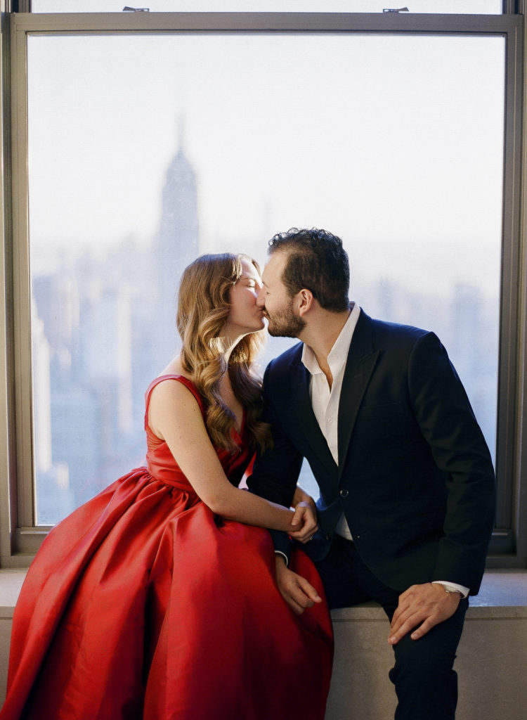 A woman in a red dress and a man in a dark suit kissing in front of a window at Top of the Rock in NYC with the Empire State Building in the background during their engagement session with You Look Lovely Photography