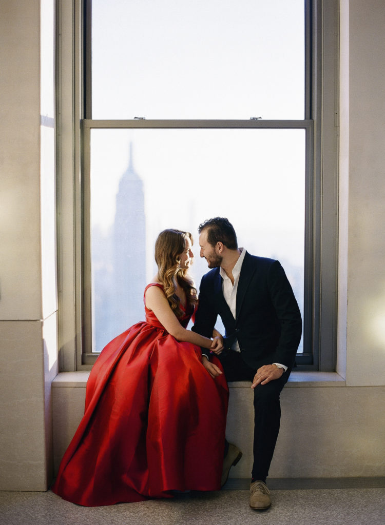 A woman in a red dress and a man in a dark suit looking into each other's eyes in front of a window at Top of the Rock in NYC with the Empire State Building in the background during their engagement session with You Look Lovely Photography