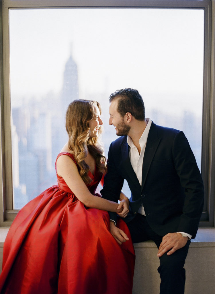 A woman in a red dress and a man in a dark suit looking into each other's eyes in front of a window at Top of the Rock in NYC with the Empire State Building in the background during their engagement session with You Look Lovely Photography