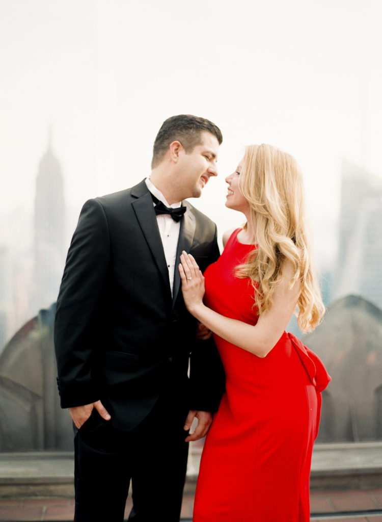 Top of the Rock Observatory Deck engagement photo in NYC of a dark-haired man in a dark suit and bowtie and a blonde woman in a red dress looking into each other's eyes., image by You Look Lovely Photography