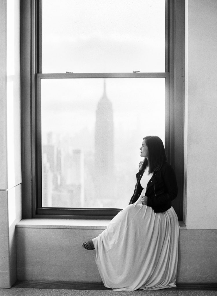 A black and white photo of a woman in a leather jacket and white dress sitting in front of a window at Top of the Rock in NYC with the Empire State Building in the background, photographed by You Look Lovely Photography