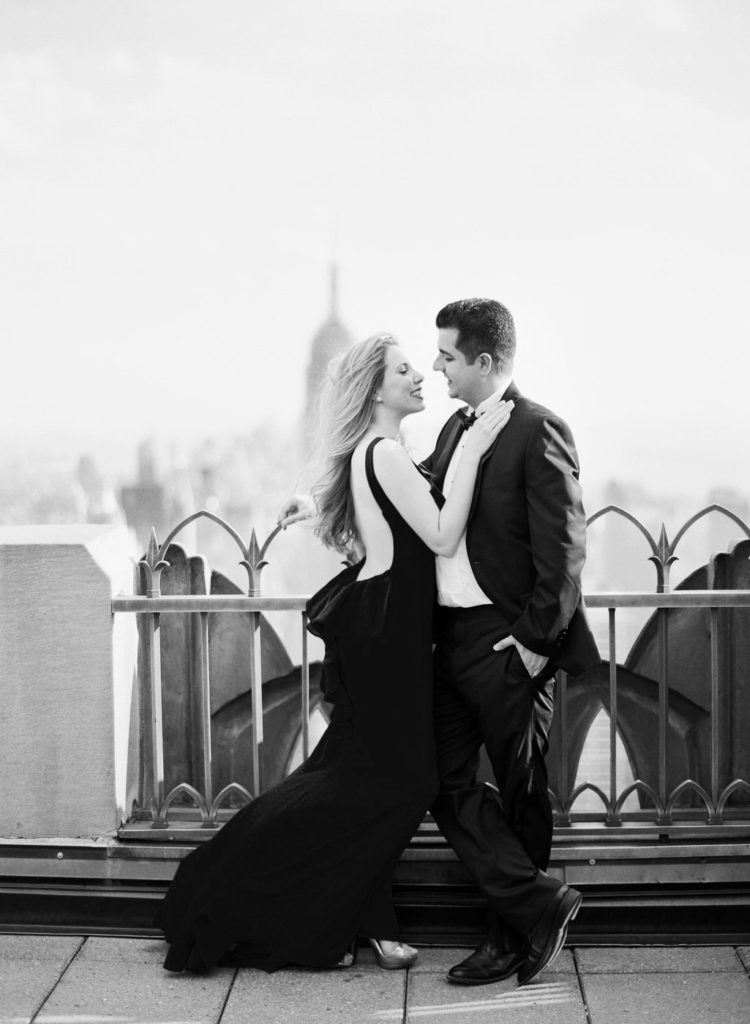 Black and white Top of the Rock engagement photo of a woman in a dress with her hand on her fiance's shoulder with the Empire State Building in the distance, image by You Look Lovely Photography