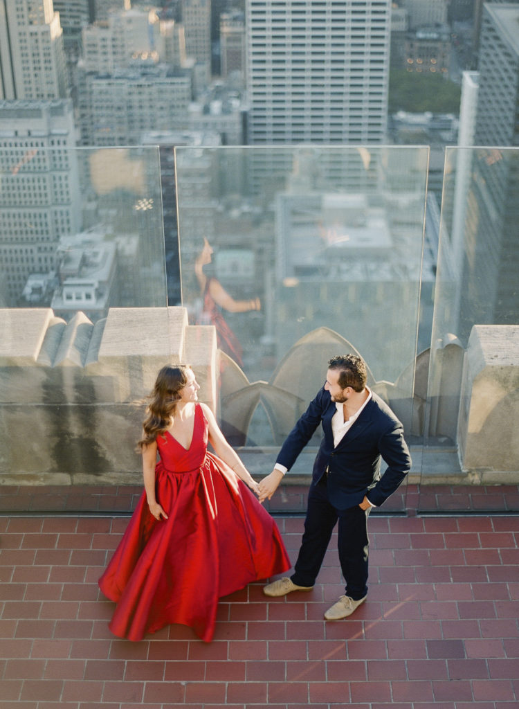 Top of the Rock engagement photo of a woman in a red dress and a man in a dark suit holding hands and taken from above with NYC buildings in the background, image by You Look Lovely Photography