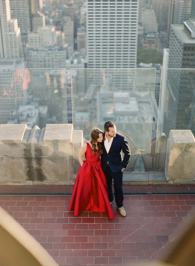 A sophisticated couple with a woman in a fancy red dress and a man in a dark tailored suit standing together on the rooftop observatory deck of Top of the Rock with NYC Midtown behind them during their engagement photo session, image by You Look Lovely Photography