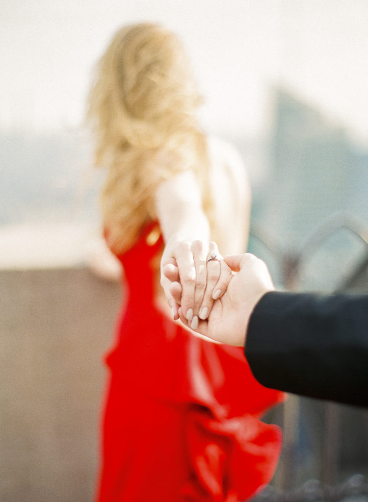 A blonde woman in a red dress reaching her arm back to take hold of her fiance's hand during their engagement photos at Top of the Rock NYC, image by You Look Lovely Photography