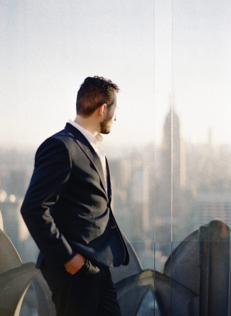 A sophisticated man standing with hands in pockets on Top of the Rock rooftop observatory deck looking out toward the Empire State Building during an engagement photo session, image by You Look Lovely Photography