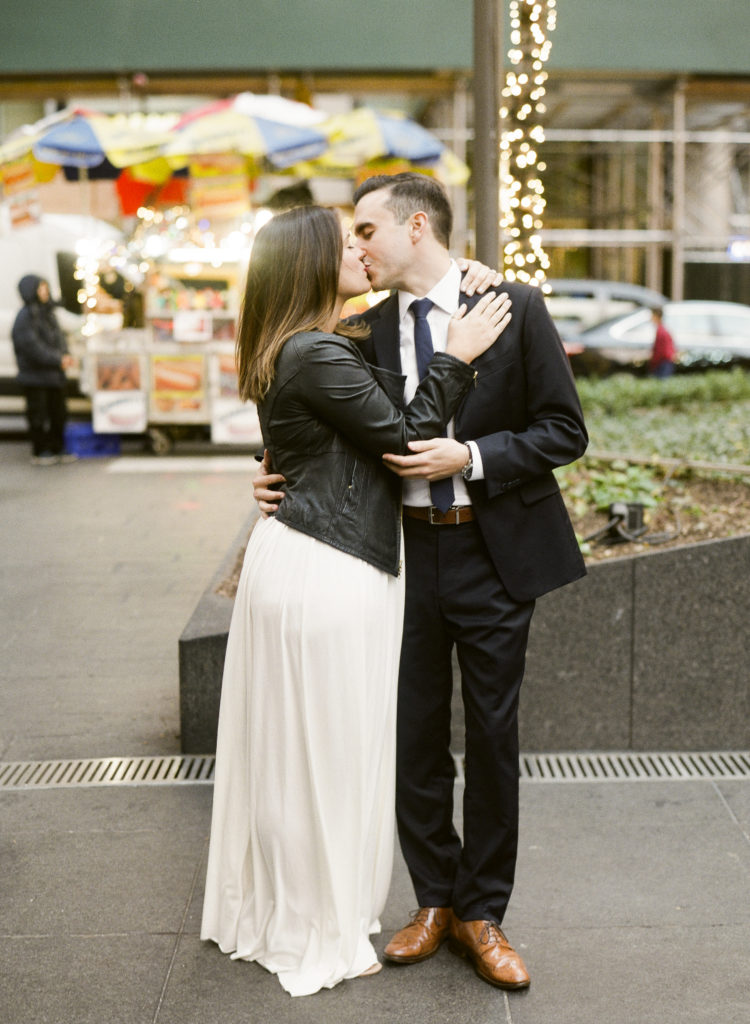 A couple kissing on a New York City street in front of a hot dog cart during their NYC engagement session, the woman is wearing a leather jacket over a white dress and her fiance is wearing a black suit and brown shoes, image by You Look Lovely Photography