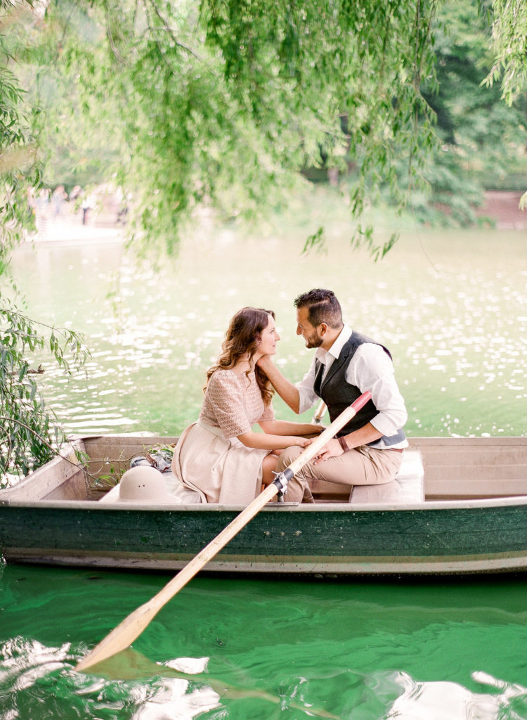 A man lightly touching his fiancee in a rowboat beneath a weeping willow tree in the lake at Central Park, during New York City engagement photos taken by You Look Lovely Photography