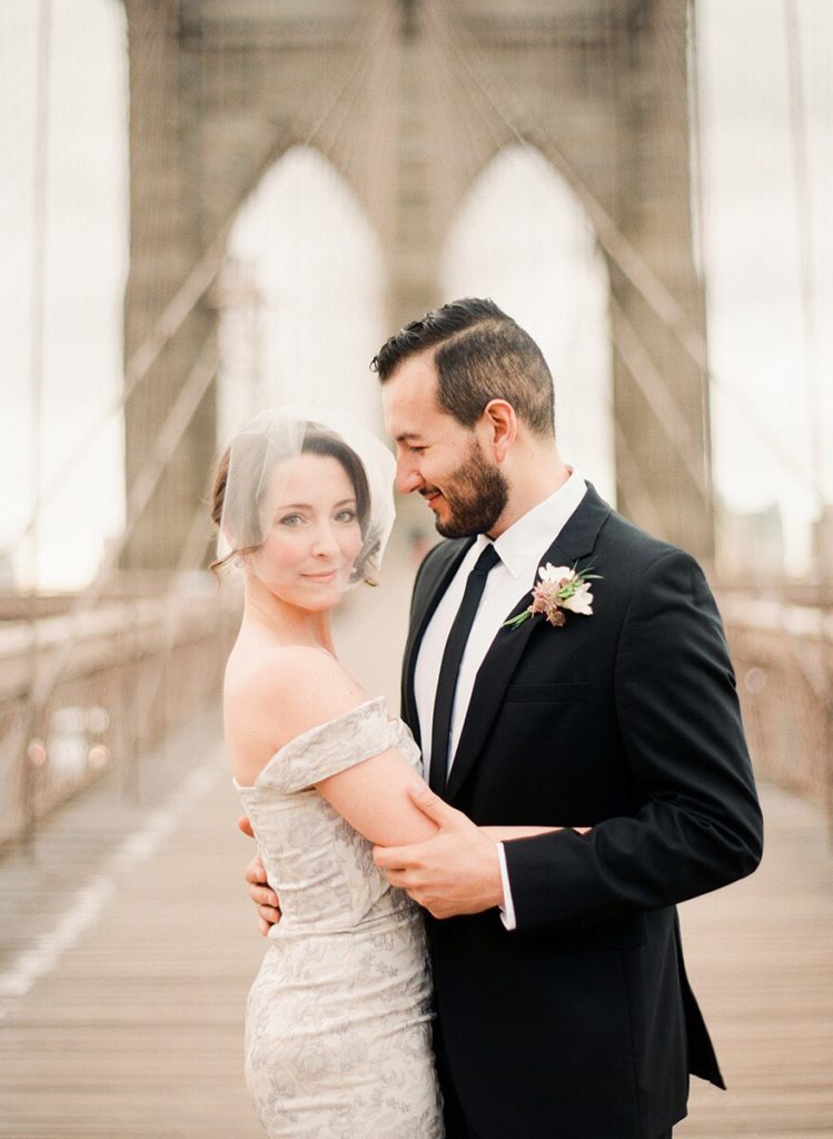 A woman in a veil and white dress and a man in a suit holding each other close on the Brooklyn Bridge during an elopement photoshoot by You Look Lovely Photography