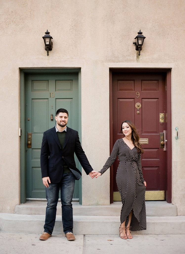 A couple standing and holding hands in front of a green and red set of doors on Washington Mews during a romantic engagement photoshoot in NYC.