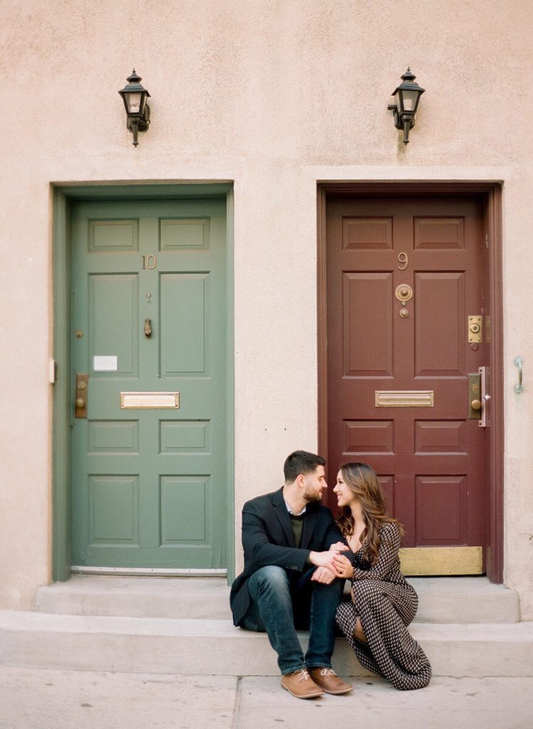 A couple sitting and holding hands in front of a green and red set of doors on Washington Mews during a romantic engagement photoshoot in NYC.