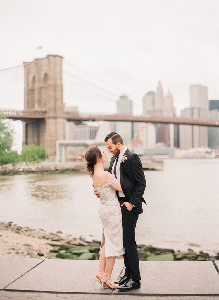 Romantic couple looking into each other's eyes at Brooklyn Bridge Park with the bridge and city behind them during NYC engagement photos by You Look Lovely Photography