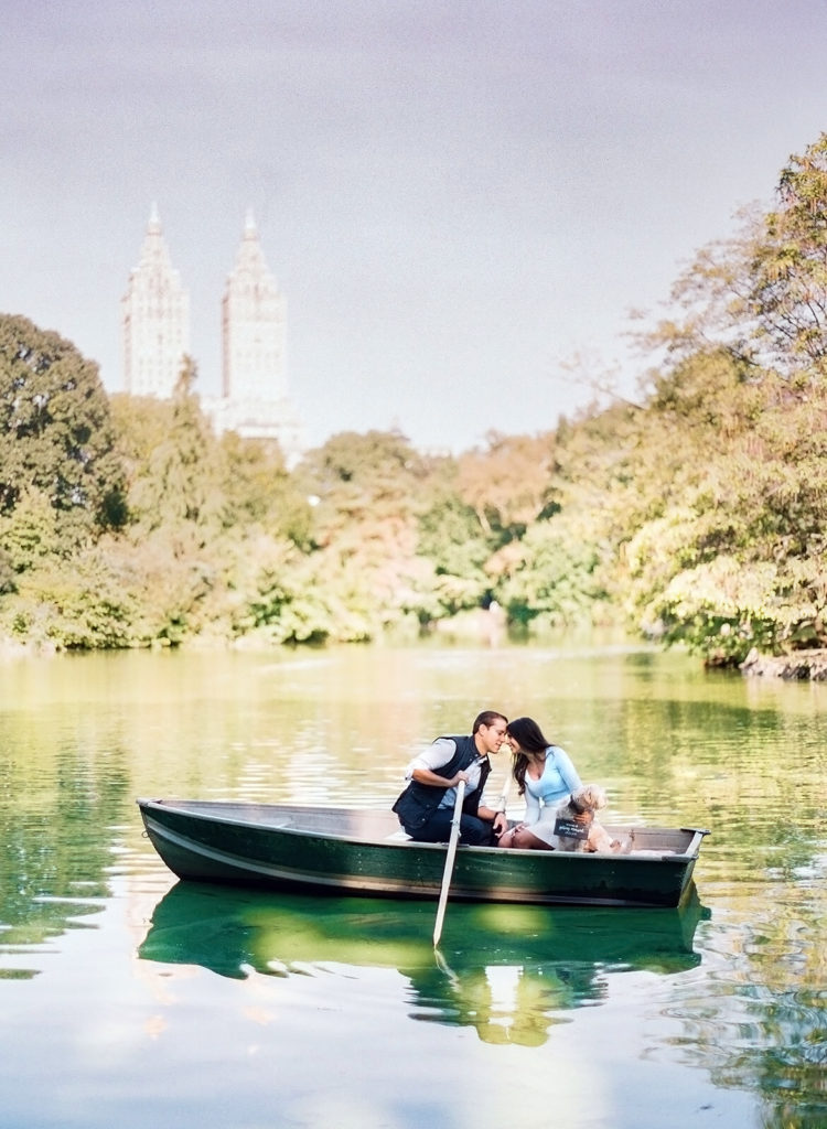 A couple and their puppy on a rowboat on the lake at Central Park with the two towers of The Eldorado building in the distance during NYC engagement photos by You Look Lovely Photography