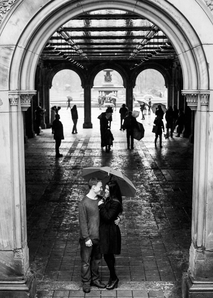 A couple standing under an arch at Bethesda Terrace Arcade with an umbrella in the rain and Bethesda Fountain in the background photographed by You Look Lovely Photography