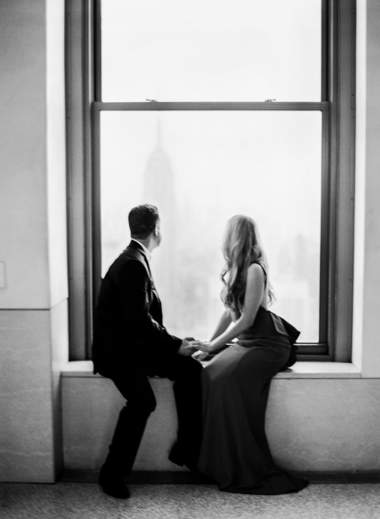 A black and white image of a couple looking out a window at the Empire State Building from the Top of the Rock during NYC engagement photos by You Look Lovely Photography
