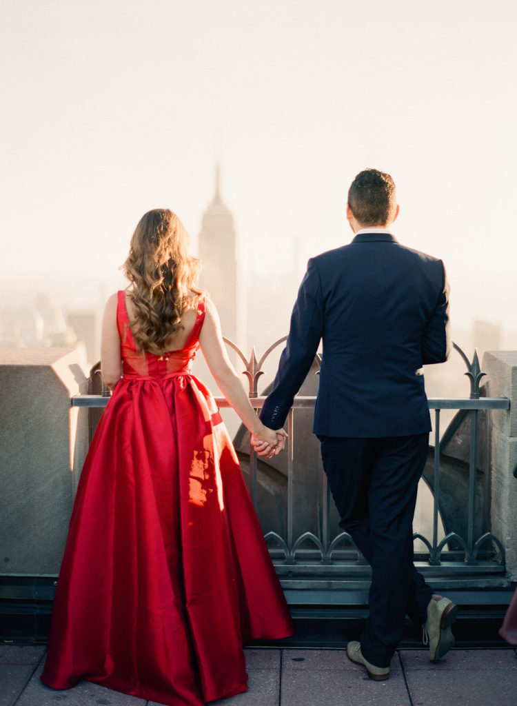 A woman and her fiancé standing on the rooftop deck at Top of the Rock, gazing at the Empire State Building in the distance during romantic NYC engagement photos by You Look Lovely Photography