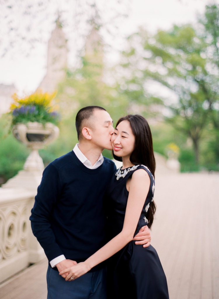 A woman smiling as her fiance kisses her cheek in front of Bow Bridge in Central Park, NYC, during engagement photos in NYC by You Look Lovely Photography