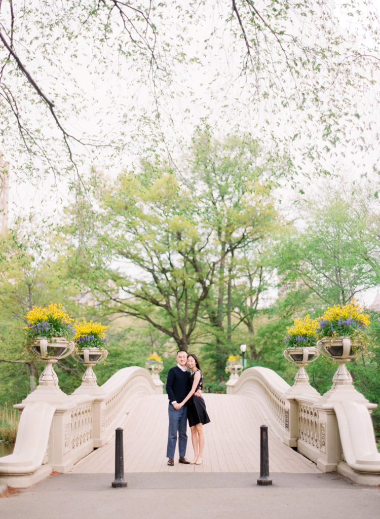 A young couple holding each other at the foot of Bow Bridge in Central Park during NYC engagement photosn by You Look Lovely Photography with yellow and purple flowers around them