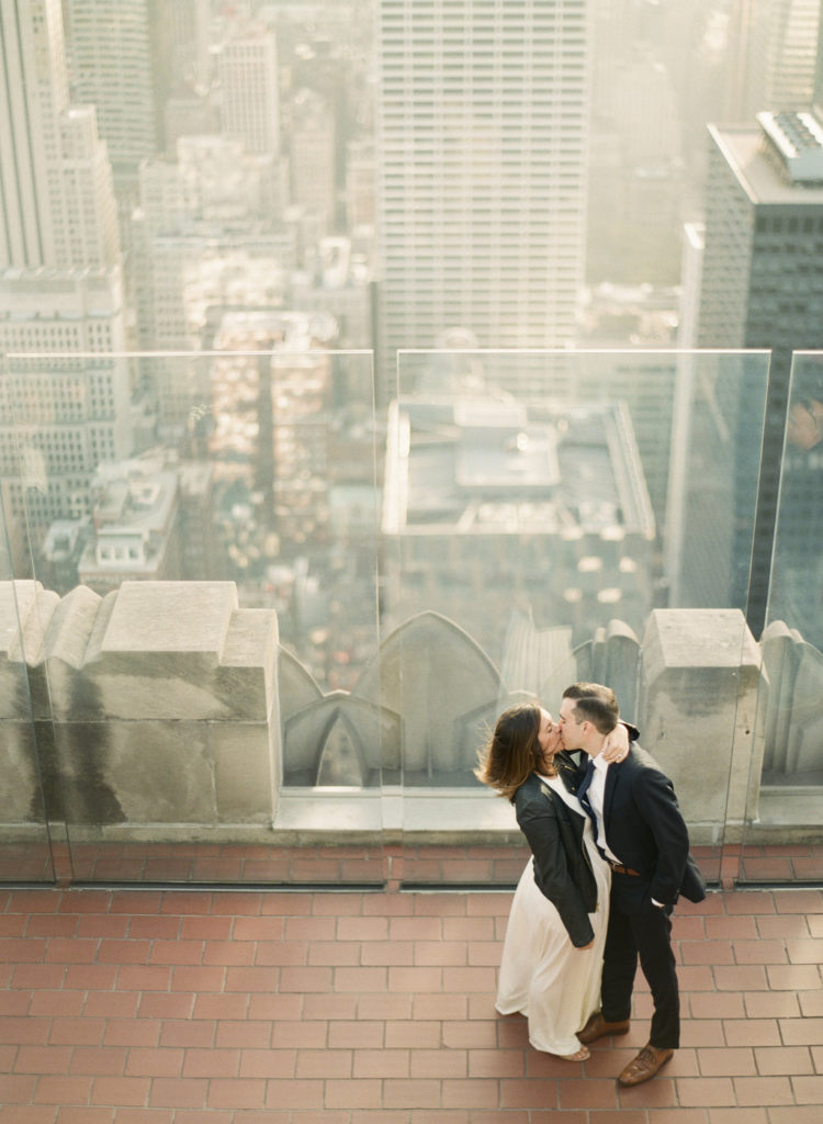 A man in a suit kissing his fiancee on the rooftop deck at Top of the Rock during romantic engagement pictures in NYC with buildings in the distance photographed by You Look Lovely Photography