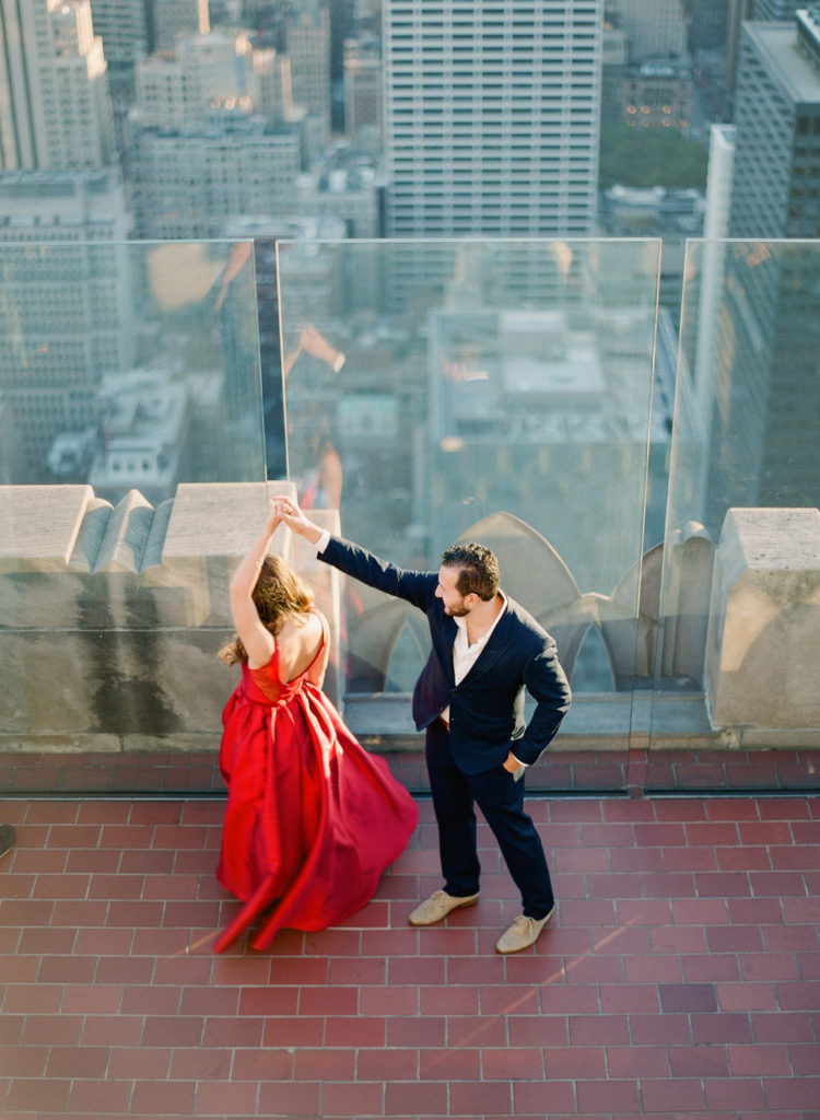 A man in a dark suit twirling a woman in a red dress during their roof top engagement photo session at the Top of the Rock in NYC with You Look Lovely Photography