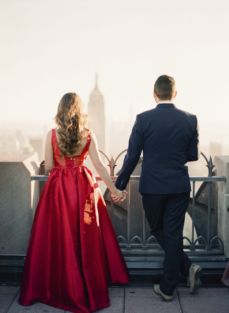 A woman in a red dress and a man in a dark suit standing with their backs toward the camera and holding hands as they look off toward the Empire State Building in the distance during their engagement photos at Top of the Rock in NYC, image by You Look Lovely Photography