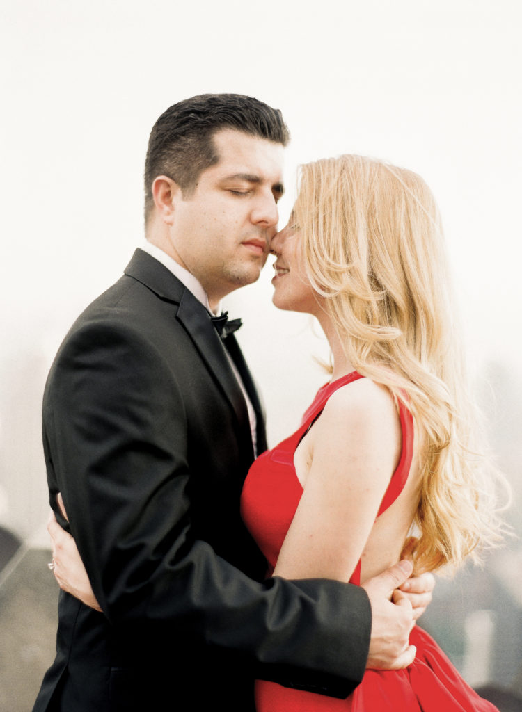 A romantic moment between a man with dark hair wearing a black suit and bowtie and a blonde woman in a red dress during their engagement photos at Top of the Rock in NYC with You Look Lovely Photography