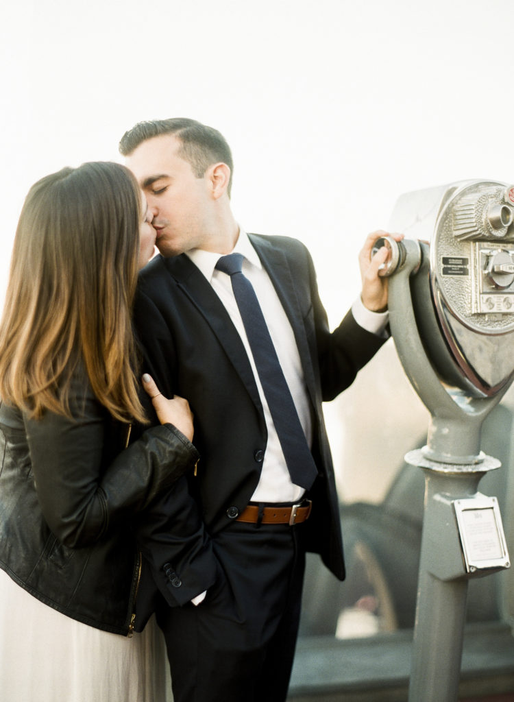 An engaged couple kissing with their eyes closed at Top of the Rock by coin-operated binoculars with a woman in a leather jacket and white dress and a man in a black suit and necktie during a photo shoot with You Look Lovely Photography
