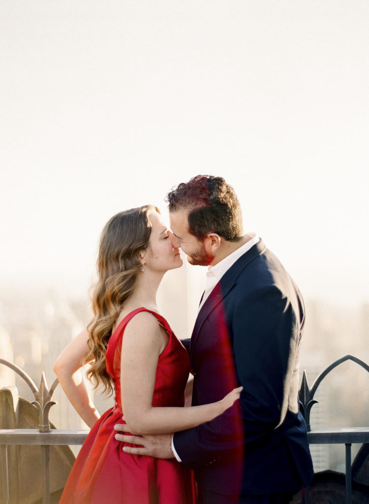 A woman in a red dress and a man in a dark suit about to kiss during their rooftop engagement photo shoot in NYC at Top of the Rock, image by You Look Lovely Photography