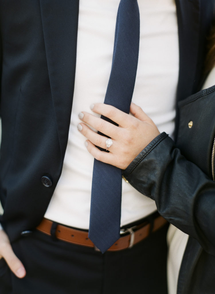 A woman's hand delicately holding her fiance's navy blue necktie and showing off her engagement ring during a couples portrait session in NYC at Top of the Rock, image by You Look Lovely Photography