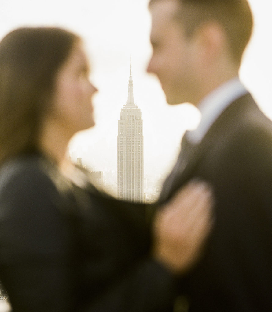 An artistic image of the Empire State Building framed by a blurred out couple looking into each others eyes during their engagement photos with You Look Lovely Photography