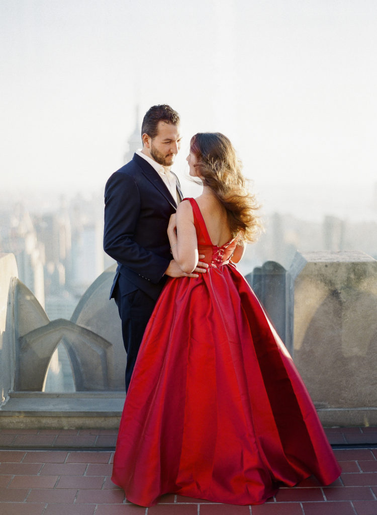 A elegantly dressed couple looking at each other as the wind blows during their The Top of the Rock engagement photos with You Look Lovely Photography