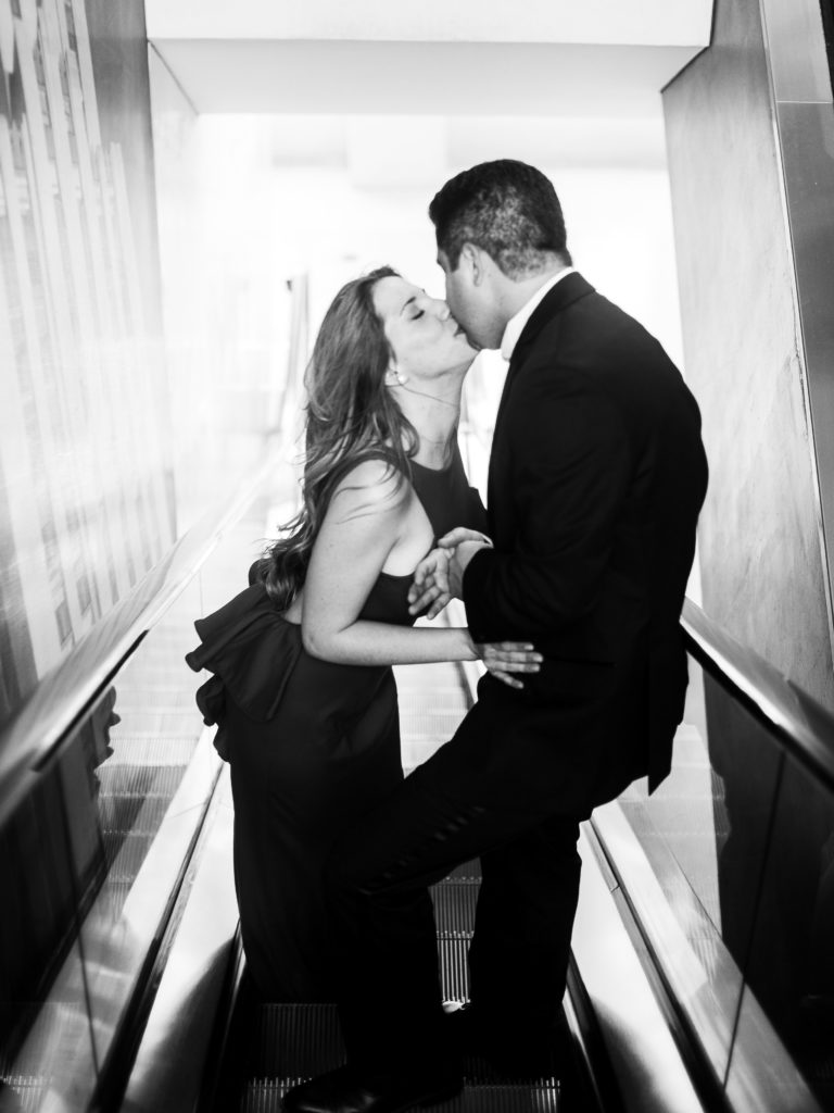 A black and white photo of a newly engaged couple kiss while going down the escalator at Top of the Rock during their engagement photos with You Look Lovely Photography