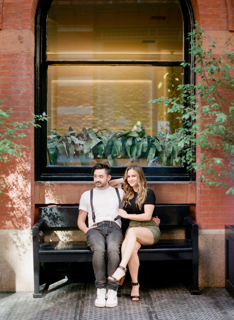 Couple sits on a bench nestled into a brick wall in SoHo NYC for their editorial engagement photos with You Look Lovely Photography