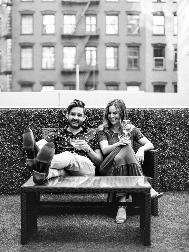 A couple laughs while sharing a drink on a rooftop overlooking historic SoHo buildings during their editorial style engagement photos with You Look Lovely Photography