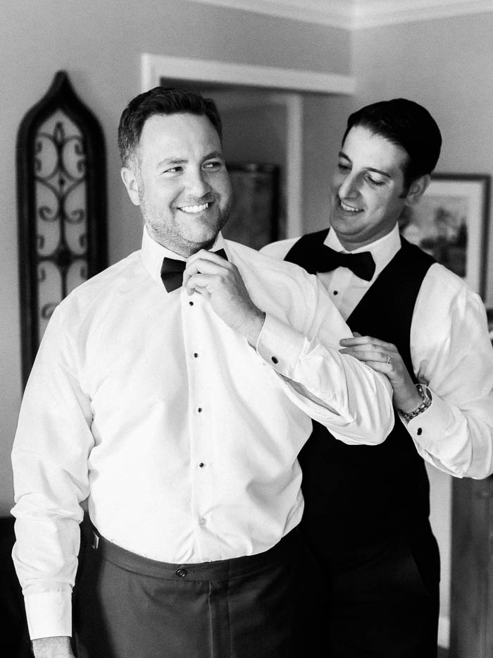 A black and white image of a groom getting ready and his best man helping him with his tie photographed by You Look Lovely Photography