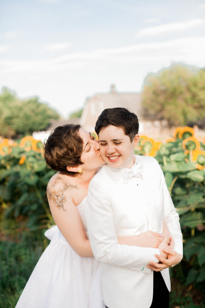 Sweet couple embrace with a sunflower field behind them during their same-sex wedding at the Queens County Farm photographed by You Look Lovely Photography
