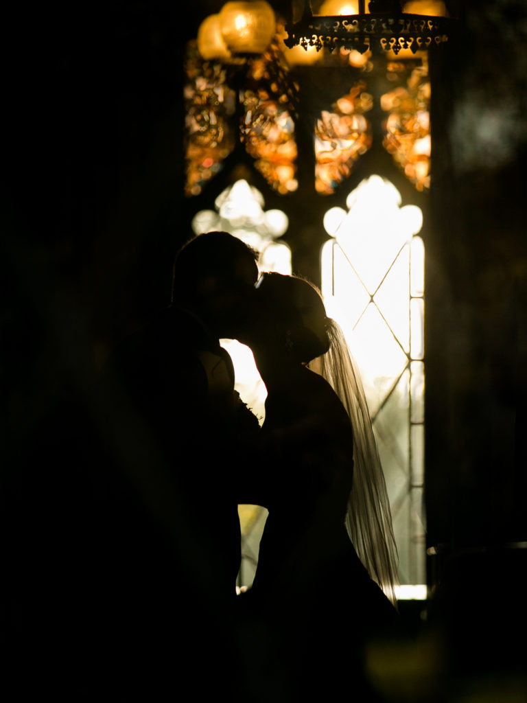 Silhouette of bride and groom kissing with stained glass windows of the Lyndhurst Castle in the background photographed by You Look Lovely Photography