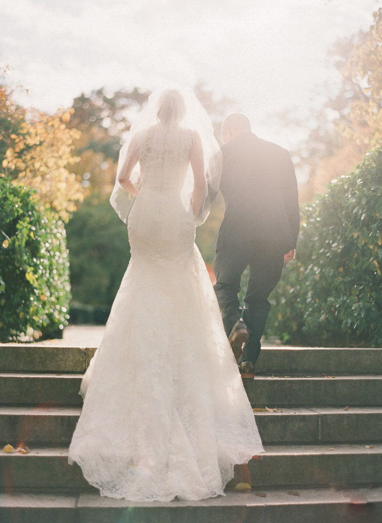 An image of a bride and her father from behind as they walk towards the aisle at the Central Park Conservatory Gardens photographed by You Look Lovely Photography